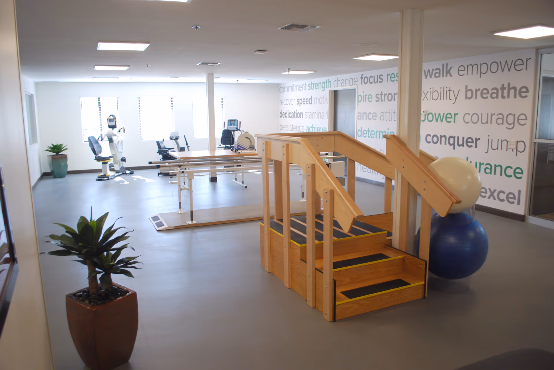 A spacious physical therapy or rehabilitation room with exercise equipment including stationary bikes, parallel bars, and a wooden staircase for mobility training. The room has large windows letting in natural light, motivational words on the wall, and potted plants for decoration.