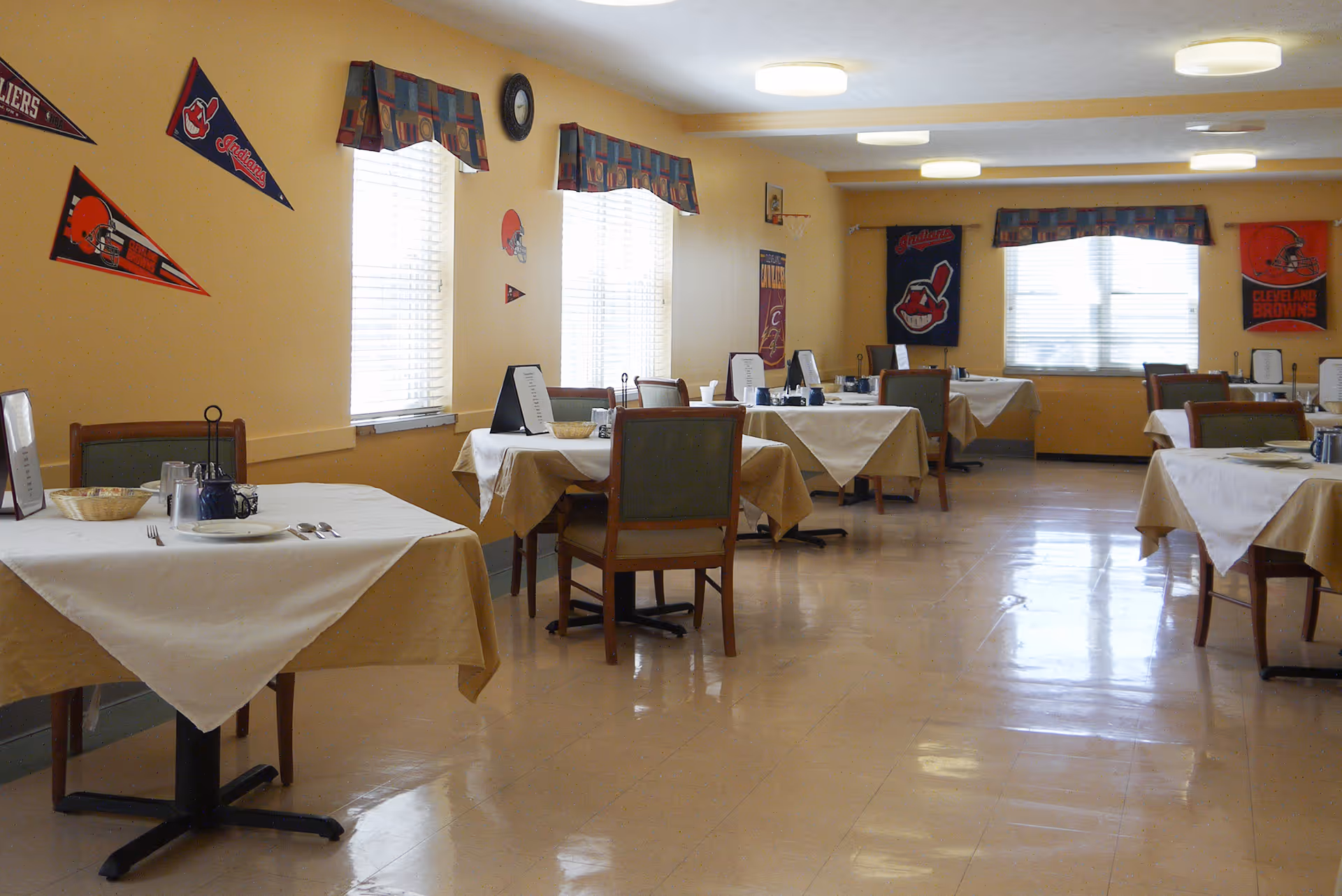 A dining room with several tables covered with beige and white tablecloths, each set with plates, utensils, and condiments. The walls are decorated with sports pennants and banners, and windows with patterned valances allow natural light to enter the room.