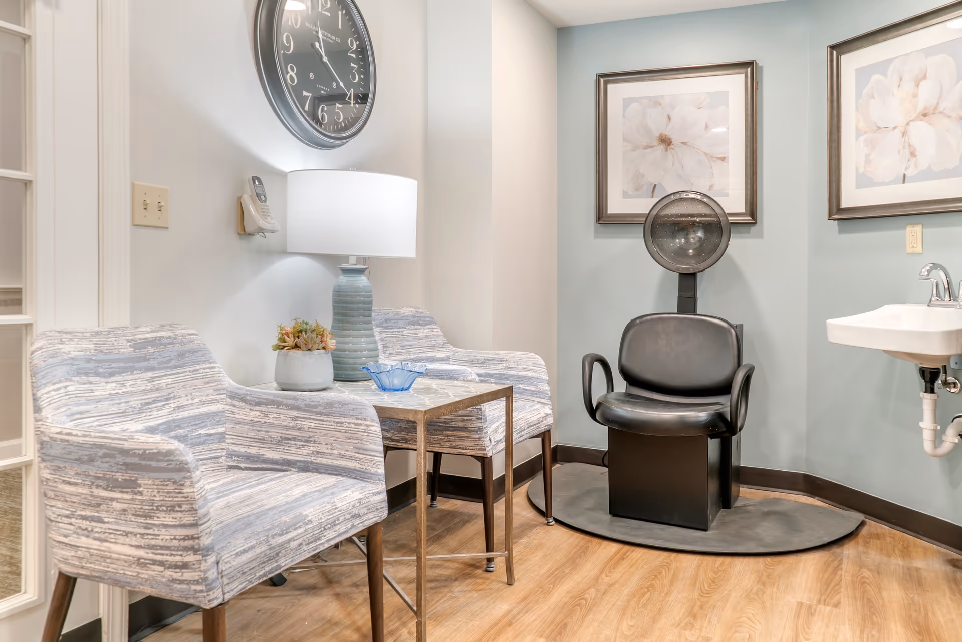 Small salon seating area with two patterned armchairs, a side table and lamp, a hooded hair dryer chair and sink against a pale blue wall.
