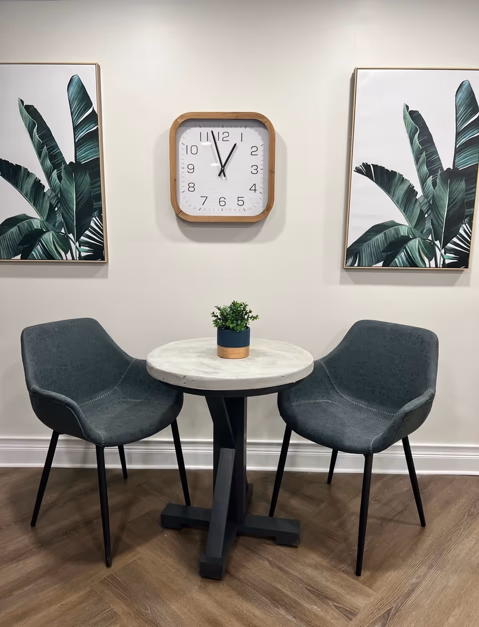 A small round table with a white marble top and black base is flanked by two dark gray upholstered chairs with black legs. On the table is a small potted plant. Behind the table on the wall are two framed pictures of large green tropical leaves, one on each side of a square wooden-framed wall clock showing the time as 11:55.