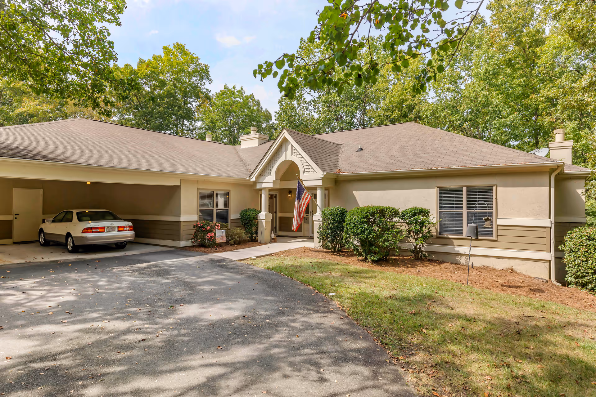 Exterior view of a single-story residential building with a carport on the left side containing a white car. The building has beige walls with brown trim, a peaked entrance with an American flag hanging, and several windows. The surrounding area has green trees and shrubs with a paved driveway in front.