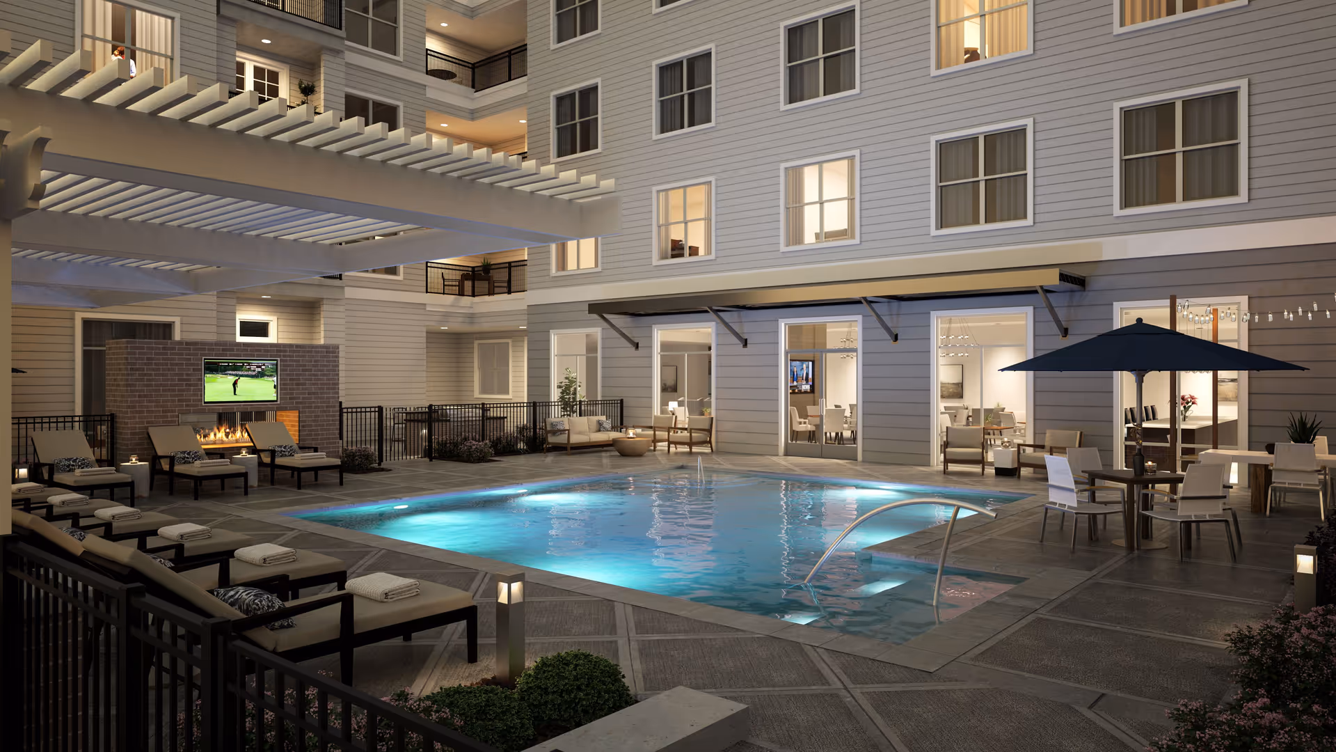 Illuminated courtyard swimming pool at night surrounded by lounge chairs, seating areas, and a multi-story building with windows.