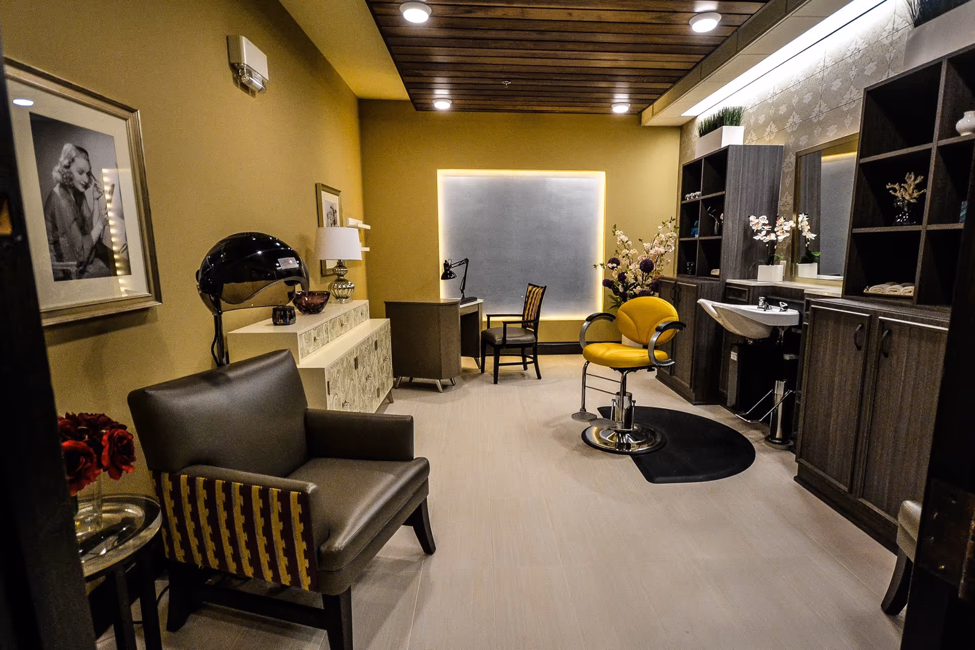 Interior view of a salon room in a senior living facility with a yellow salon chair in front of a sink, dark wood cabinets with shelves, a desk with a chair, a gray armchair, a hair dryer, and decorative flowers and framed pictures on the walls.