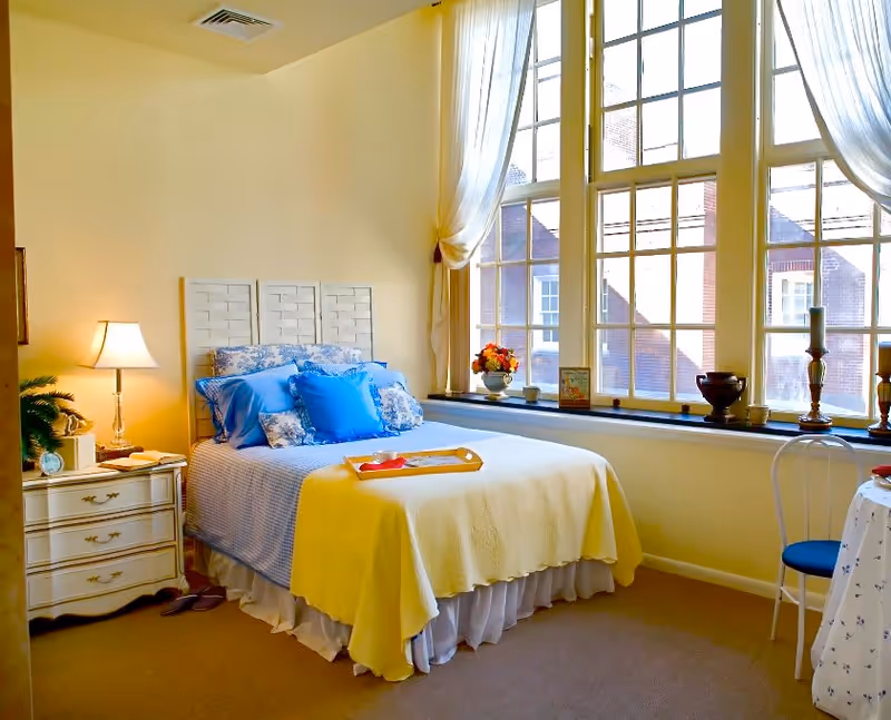 A bright bedroom with a large window letting in natural light. The bed is neatly made with blue and white bedding, and a yellow blanket draped over the foot. A bedside table with a lamp, clock, and plant is next to the bed. The window sill holds decorative items including a vase with flowers and candles. A small round table with a chair is positioned near the window.