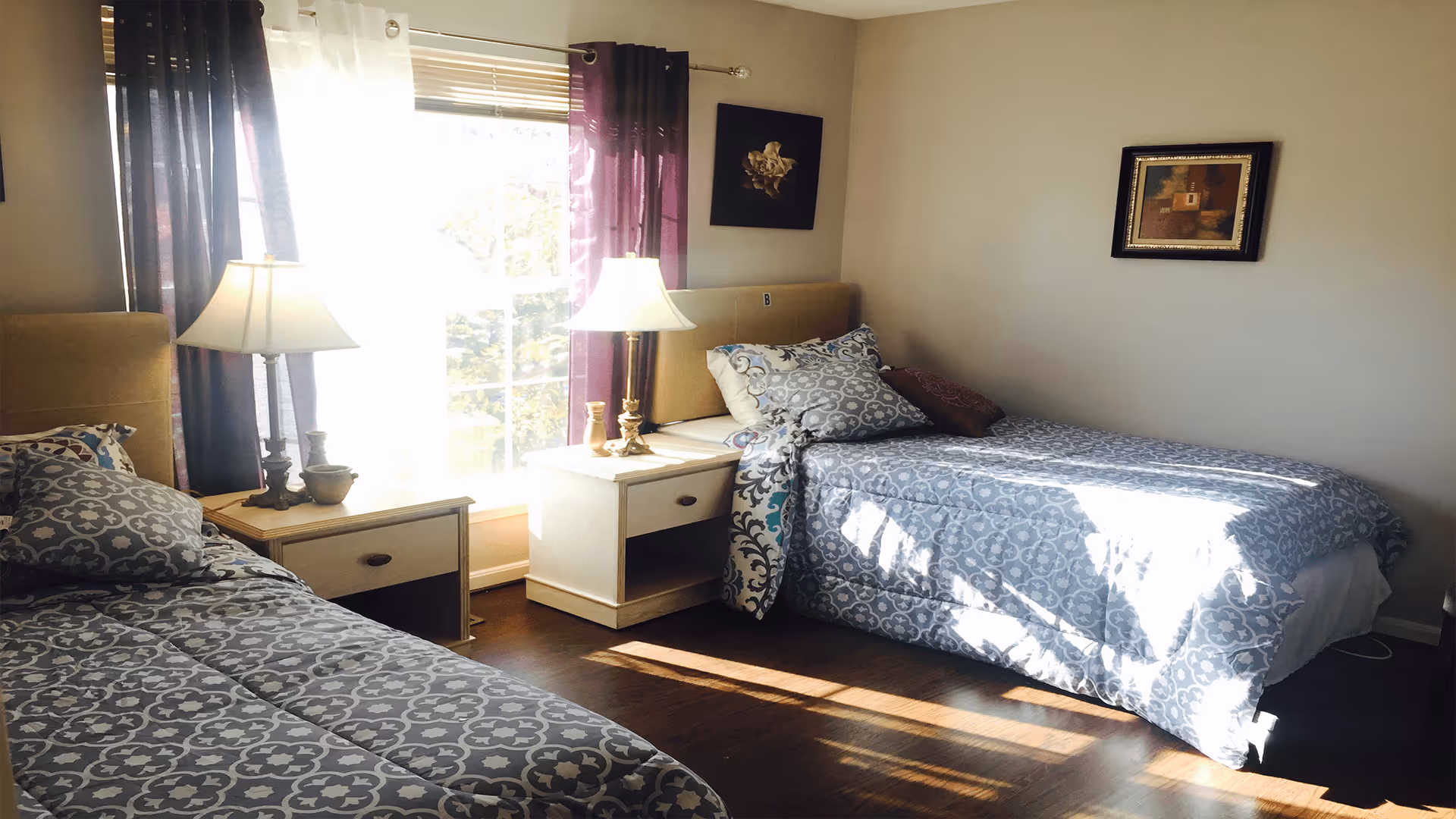 A bright bedroom with two single beds covered in matching gray and white patterned bedding. Between the beds is a small white nightstand with two lamps and decorative items. The room has dark wood flooring, light-colored walls, and a window with purple curtains letting in natural sunlight. Two framed artworks hang on the walls.