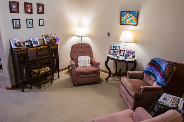 A cozy living room area with three upholstered armchairs, one with a crocheted blanket draped over it. There is a small wooden table with a lamp and framed photos, and a wooden desk with two chairs displaying more framed photos. Several framed pictures hang on the wall above the desk.