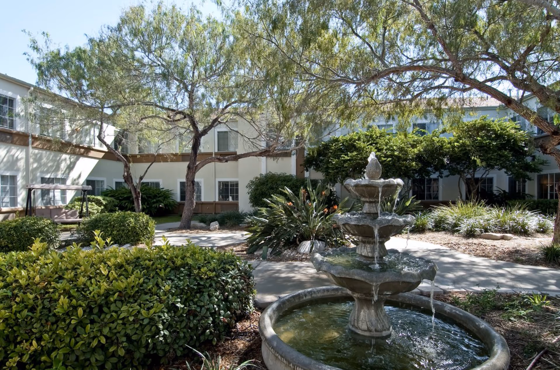 Courtyard with a tiered stone fountain surrounded by shrubs, trees, and a two-story building in the background.