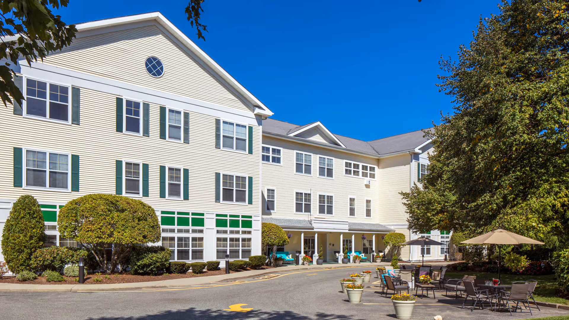 Exterior view of a three-story senior living facility building with beige siding and green window shutters under a clear blue sky. The entrance is covered with a small porch, and there are neatly trimmed bushes and trees around the building. In the foreground, there are outdoor seating areas with tables, chairs, umbrellas, and flower pots.