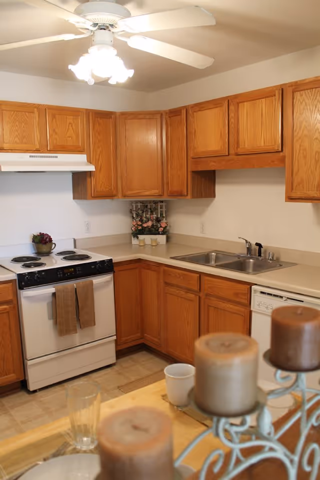 A kitchen with wooden cabinets, a white electric stove with two brown towels hanging on the handle, a double sink, and a dishwasher. There is a ceiling fan with lights above, and a small decorative plant on the countertop. In the foreground, there is a table with a glass, a white cup, and a candle holder with three brown candles.