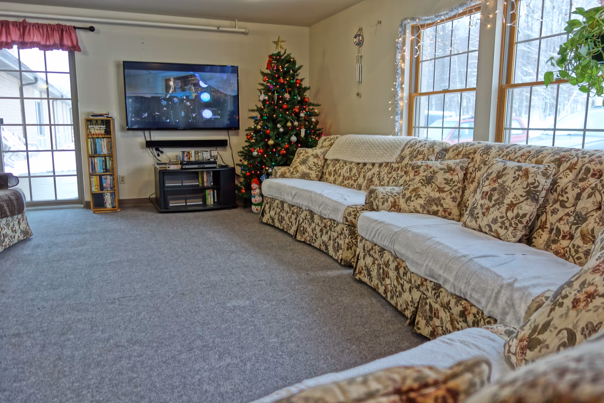 A cozy living room with floral patterned sofas covered with white throws, a decorated Christmas tree with lights and ornaments, a flat-screen TV mounted on the wall, a small entertainment center with DVDs and electronic devices, large windows with a view of snow outside, and a sliding glass door with a red valance.
