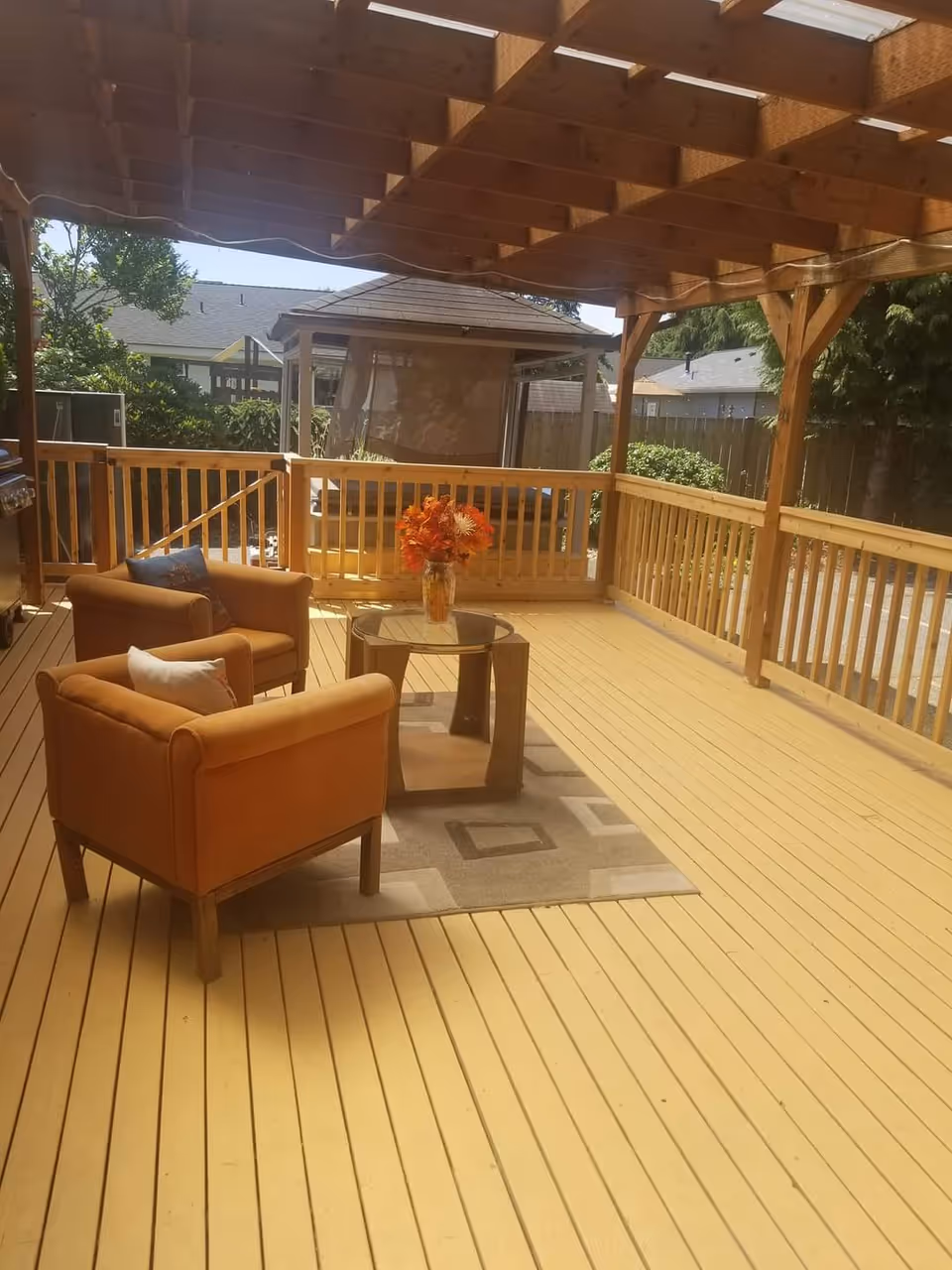 A covered outdoor wooden deck with two orange armchairs and a glass-top coffee table on a patterned rug. There is a vase with orange flowers on the table. The deck is surrounded by wooden railings and overlooks a fenced backyard with trees and neighboring houses.