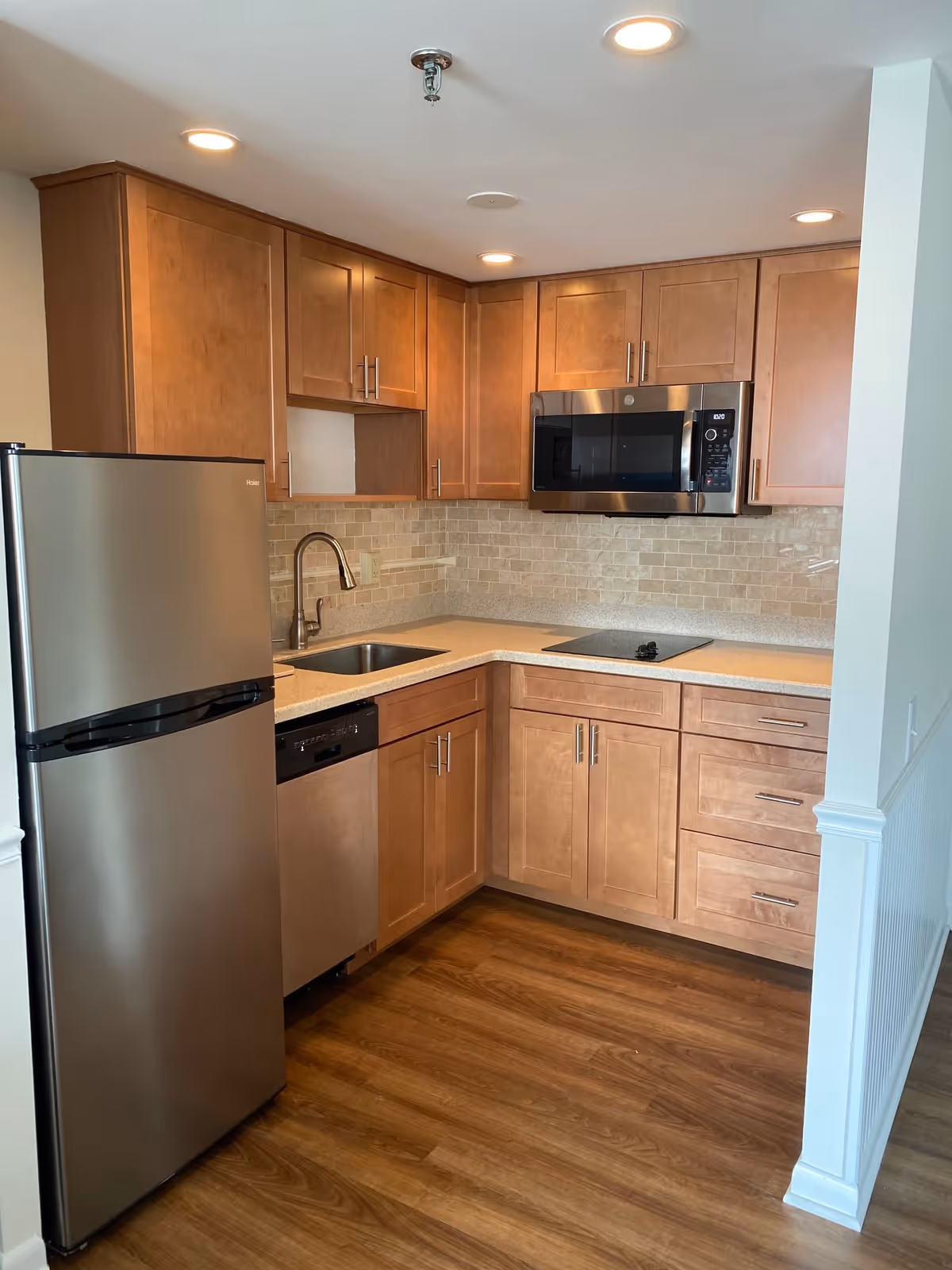 A modern kitchen with wooden cabinets, a stainless steel refrigerator, dishwasher, microwave, and a sink. The kitchen has a light-colored countertop and a tiled backsplash, with recessed ceiling lights and wood flooring.