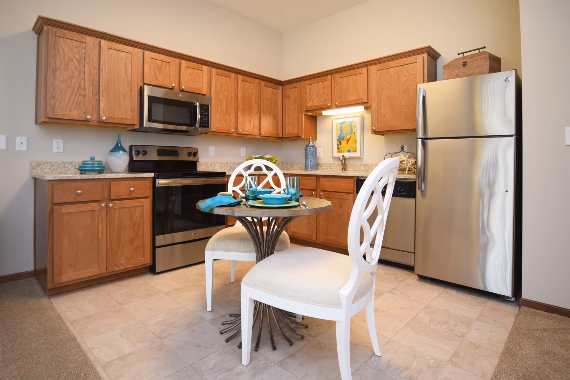 A kitchen area with wooden cabinets, stainless steel appliances including a refrigerator, stove, microwave, and dishwasher. There is a small round table with two white cushioned chairs set with turquoise plates, bowls, and glasses. The floor is tiled and the walls are painted beige.