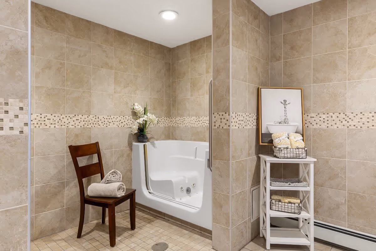 A tiled bathroom with beige walls and floor featuring a walk-in bathtub with a glass door. A wooden chair with rolled towels is placed next to the tub. A small white shelving unit holds baskets with towels and a framed picture of a giraffe in a bathtub is displayed on top. A vase with white flowers is on the corner ledge above the tub.