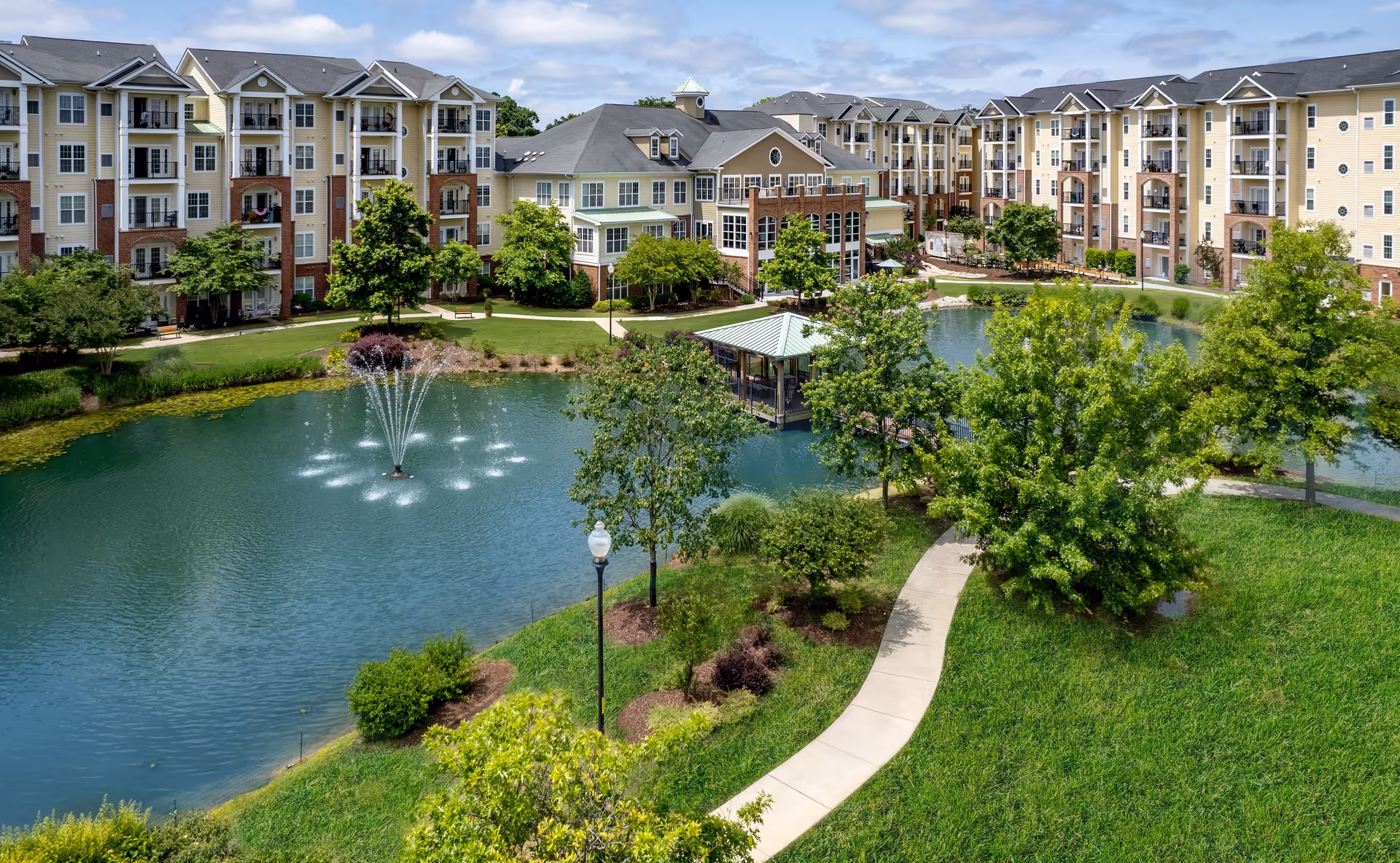 A landscaped courtyard with a pond and central fountain, gazebo, walking paths, and surrounding multi-story apartment buildings.