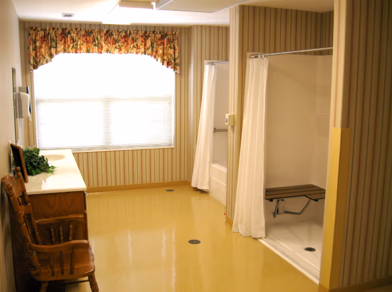 A spacious bathroom with two shower areas separated by a wall, each with a white shower curtain. One shower has a built-in bench. There is a wooden chair and a wooden vanity with a sink on the left side. The walls have vertical striped wallpaper, and a large window with floral valance lets in natural light.