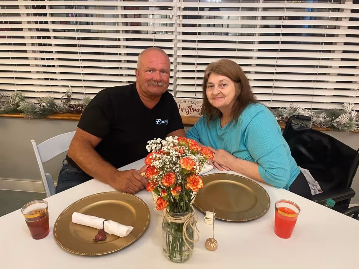 A man and a woman sitting at a dining table with gold-colored plates, napkins, and drinks. There is a vase with orange and white flowers in the center of the table. Behind them is a window with white blinds and some holiday decorations on the windowsill.