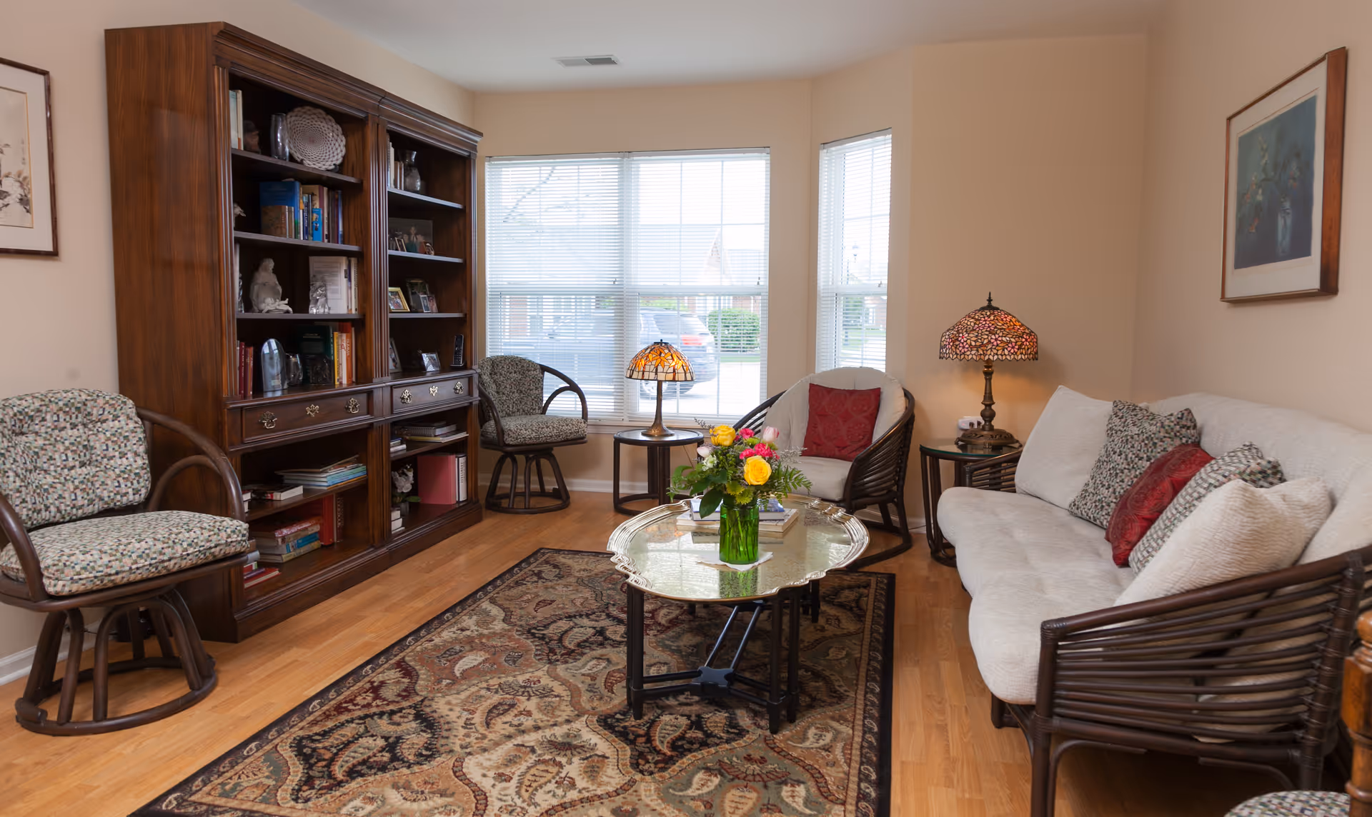 A cozy living room with a large window letting in natural light. The room features a wooden bookshelf filled with books and decorative items, two patterned cushioned chairs, a beige sofa with multiple pillows, a glass coffee table with a vase of colorful flowers, two side tables each with a Tiffany-style lamp, and a patterned area rug on a wooden floor.