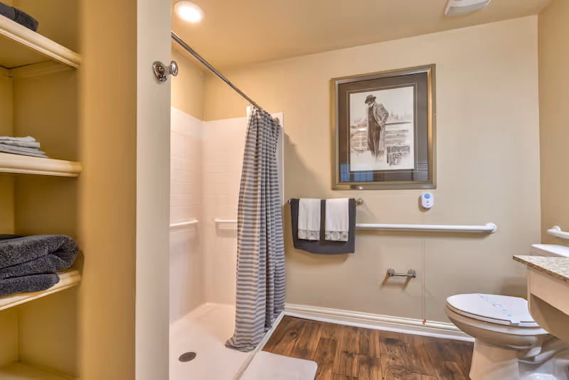 A bathroom with a walk-in shower featuring white tiles and a striped shower curtain. There are grab bars installed inside the shower and on the walls. Two folded towels hang on a towel rack beneath a framed black and white picture. A toilet with a closed lid is visible next to a countertop with a sink. Shelves with folded towels are on the left side. The floor has wood-like flooring and the walls are painted beige.