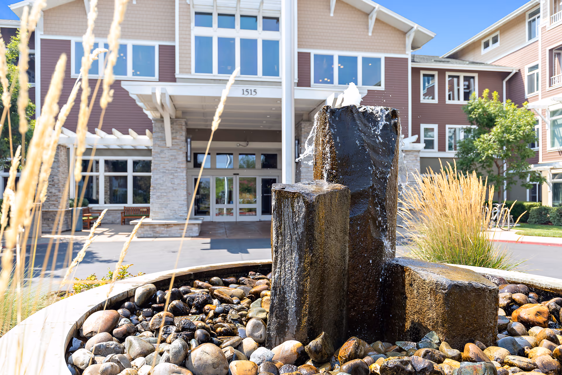 A stone water fountain with river rocks sits in front of the entrance to a multi-story retirement community building.
