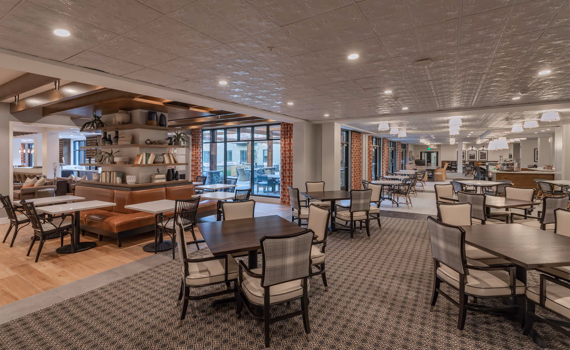 Spacious communal dining room with multiple tables and chairs, booth seating and large windows overlooking an outdoor patio.