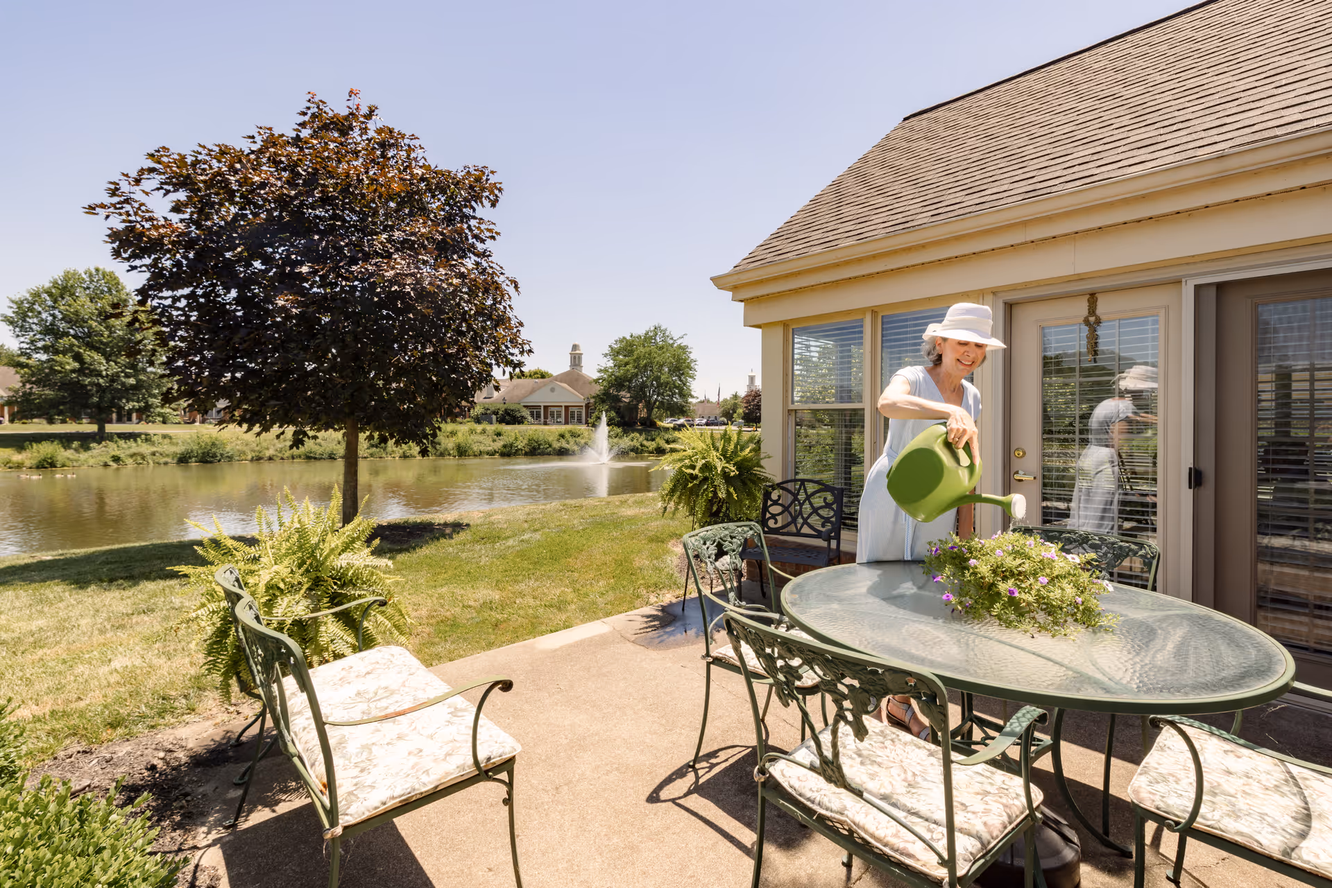 A woman wearing a white hat and light-colored dress waters a potted plant on a glass outdoor table on a patio. The patio has metal chairs with cushions and overlooks a pond with a fountain and trees under a clear blue sky.