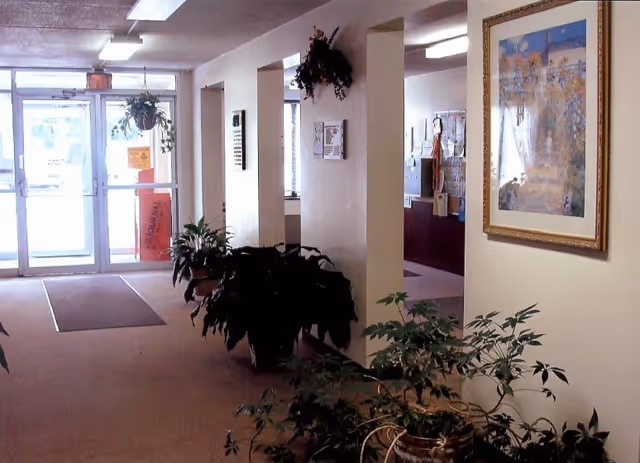 Interior view of a senior living facility hallway with beige walls and carpeted floor. Several potted plants are placed along the walls and hanging near the entrance door. A framed picture hangs on the right wall, and fluorescent ceiling lights illuminate the space. The entrance door is glass with a view of the outside.