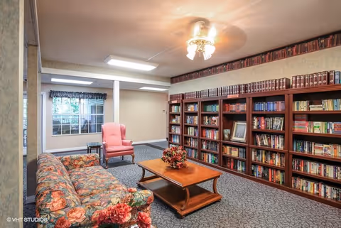 Seating area with a floral sofa, pink armchair, wooden coffee table, and a wall of bookshelves in a common room.