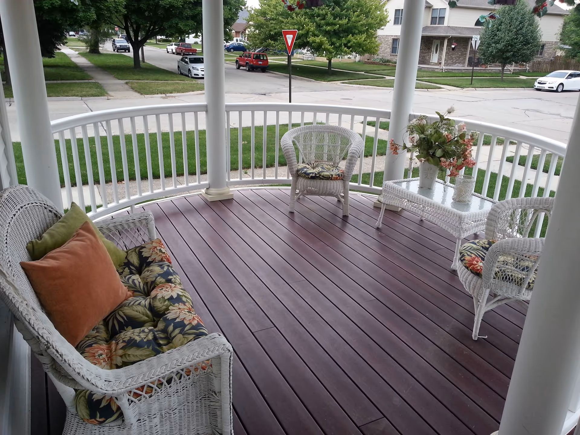 A curved porch with dark wooden flooring and white railing, furnished with white wicker chairs and a loveseat with floral cushions and pillows. A small glass-top wicker table holds a vase with flowers and a candle holder. The porch overlooks a suburban street with green lawns, trees, and parked cars.
