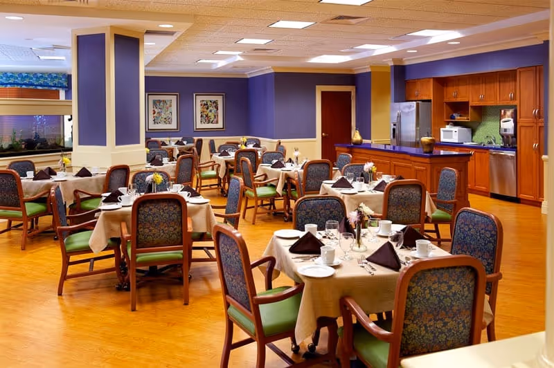 A dining room in an assisted living facility with multiple tables covered in beige tablecloths, each set with white plates, cups, silverware, and folded brown napkins. The room has wooden floors, purple and beige walls, and a kitchen area with wooden cabinets, a refrigerator, microwave, and dishwasher in the background. Several chairs with green cushions and patterned backs are arranged around the tables.