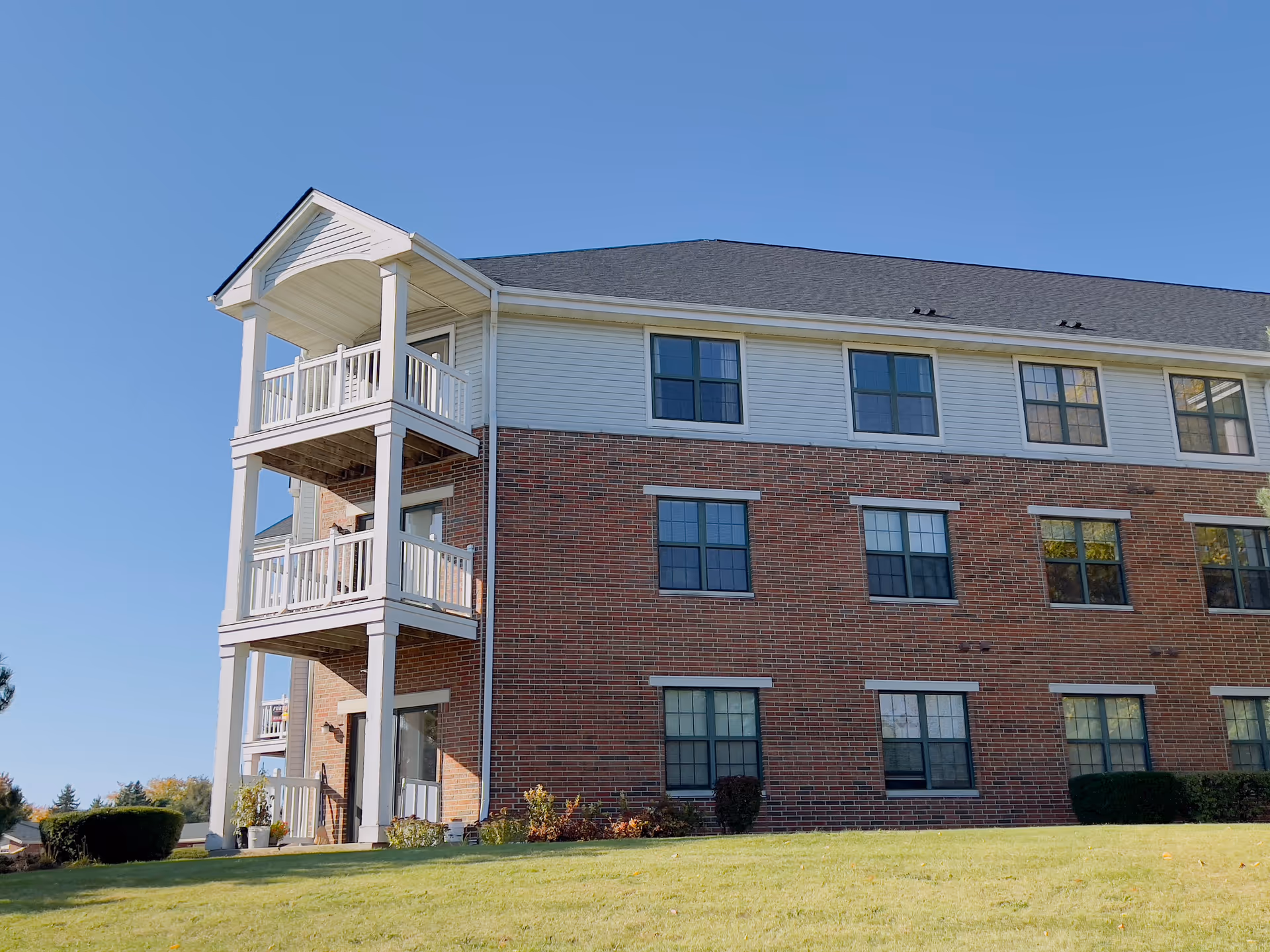 Exterior view of a three-story brick and siding apartment building with balconies on the left side, surrounded by a well-maintained grassy lawn under a clear blue sky.