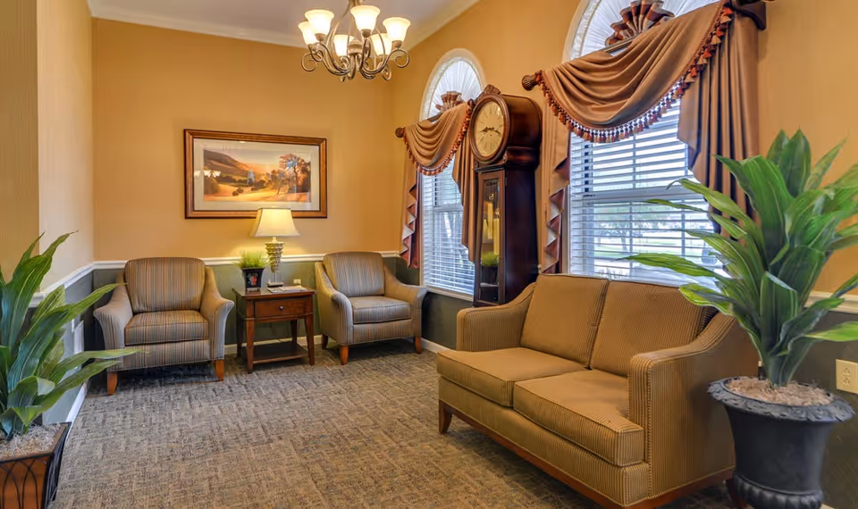 A cozy sitting area in Auburn Creek Senior Living with two striped armchairs and a small wooden side table with a lamp and plant between them. A brown couch is positioned opposite the chairs. The room features two large windows with decorative drapes, a tall grandfather clock, a framed landscape painting on the wall, and two large potted plants. The walls are painted in warm tones with white trim, and a chandelier hangs from the ceiling.