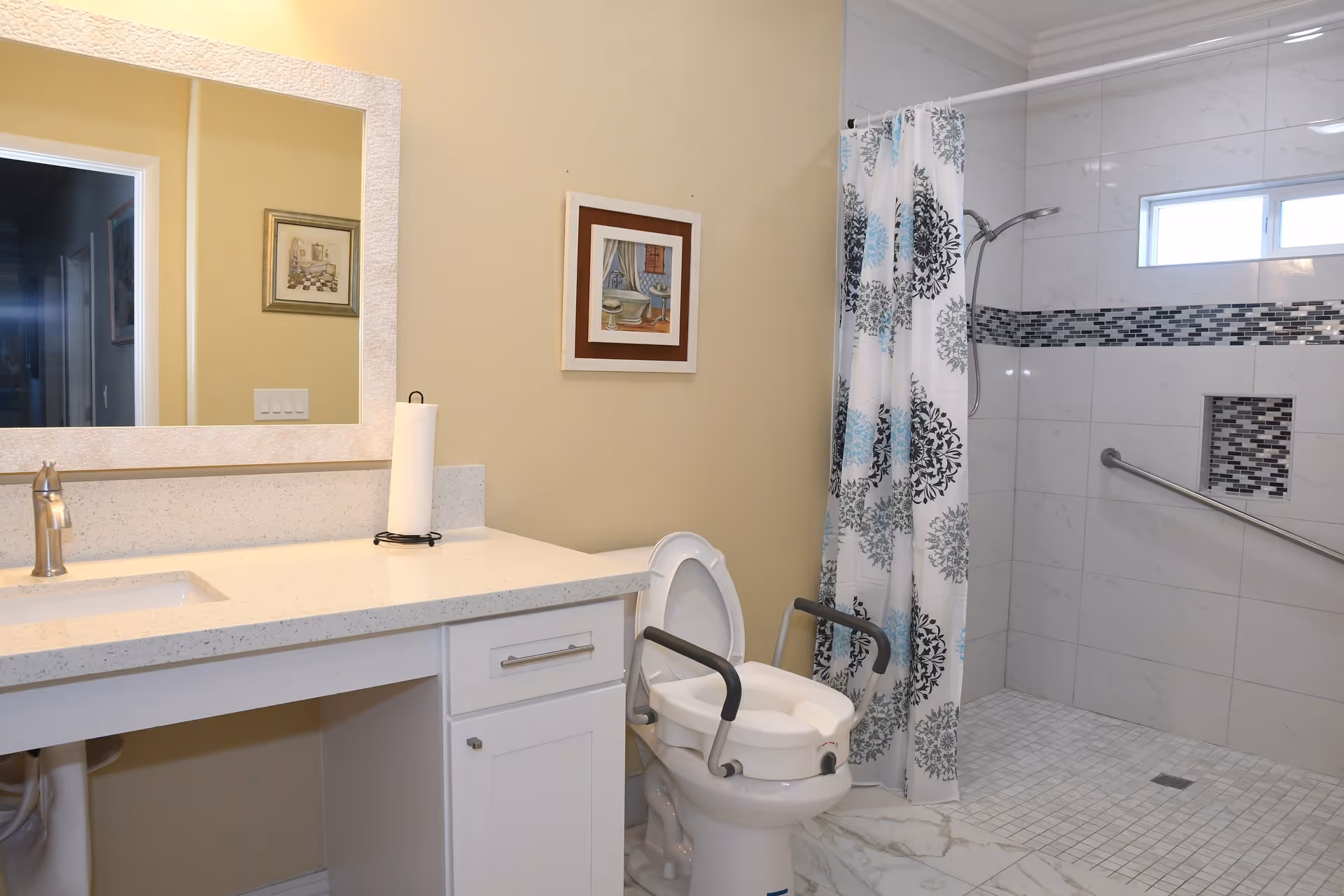 A bathroom with a white countertop and sink on the left, a toilet with safety rails in the center, and a walk-in shower with a patterned shower curtain and grab bar on the right. The walls are light-colored, and there is a framed picture hanging above the toilet.