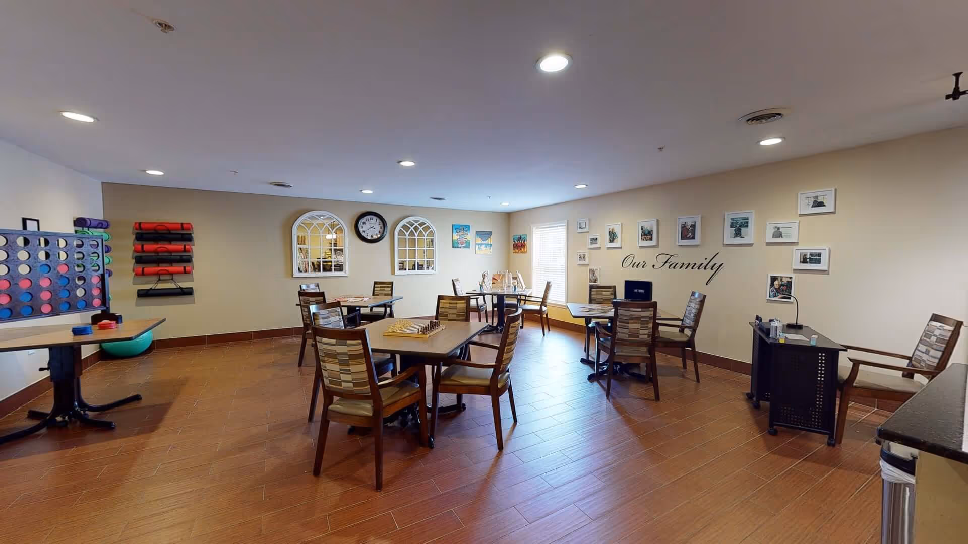 A well-lit activity room with several tables and chairs arranged for games and socializing. On the tables are board games including chess and checkers. The walls are decorated with framed photos and the words 'Our Family'. There are also two decorative mirrors and a clock on one wall. On the left side, there is a large Connect Four game and a rack holding rolled-up mats.