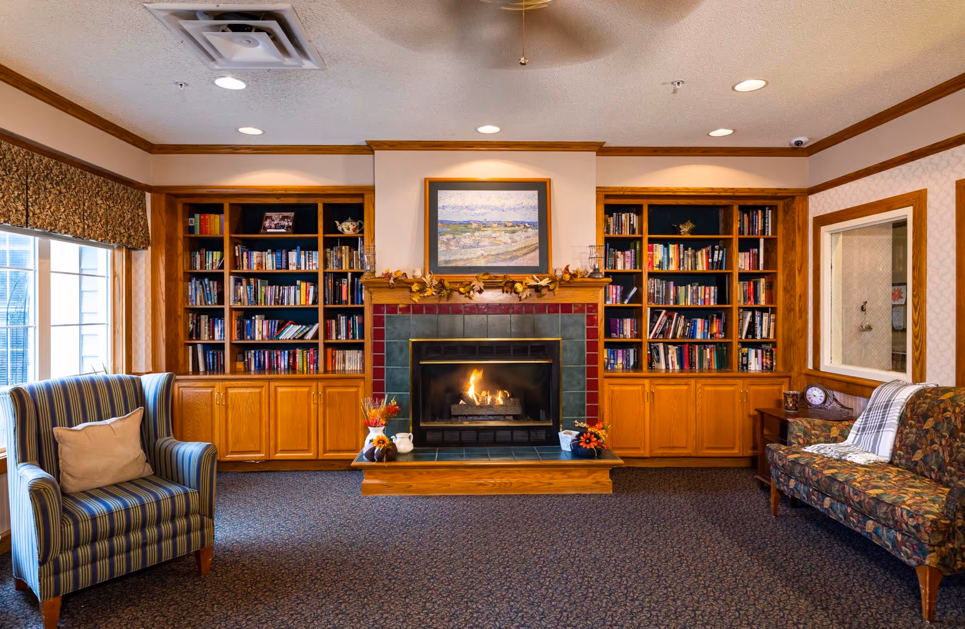 Cozy communal living room with a lit fireplace flanked by built-in bookshelves, an armchair, and a patterned sofa.