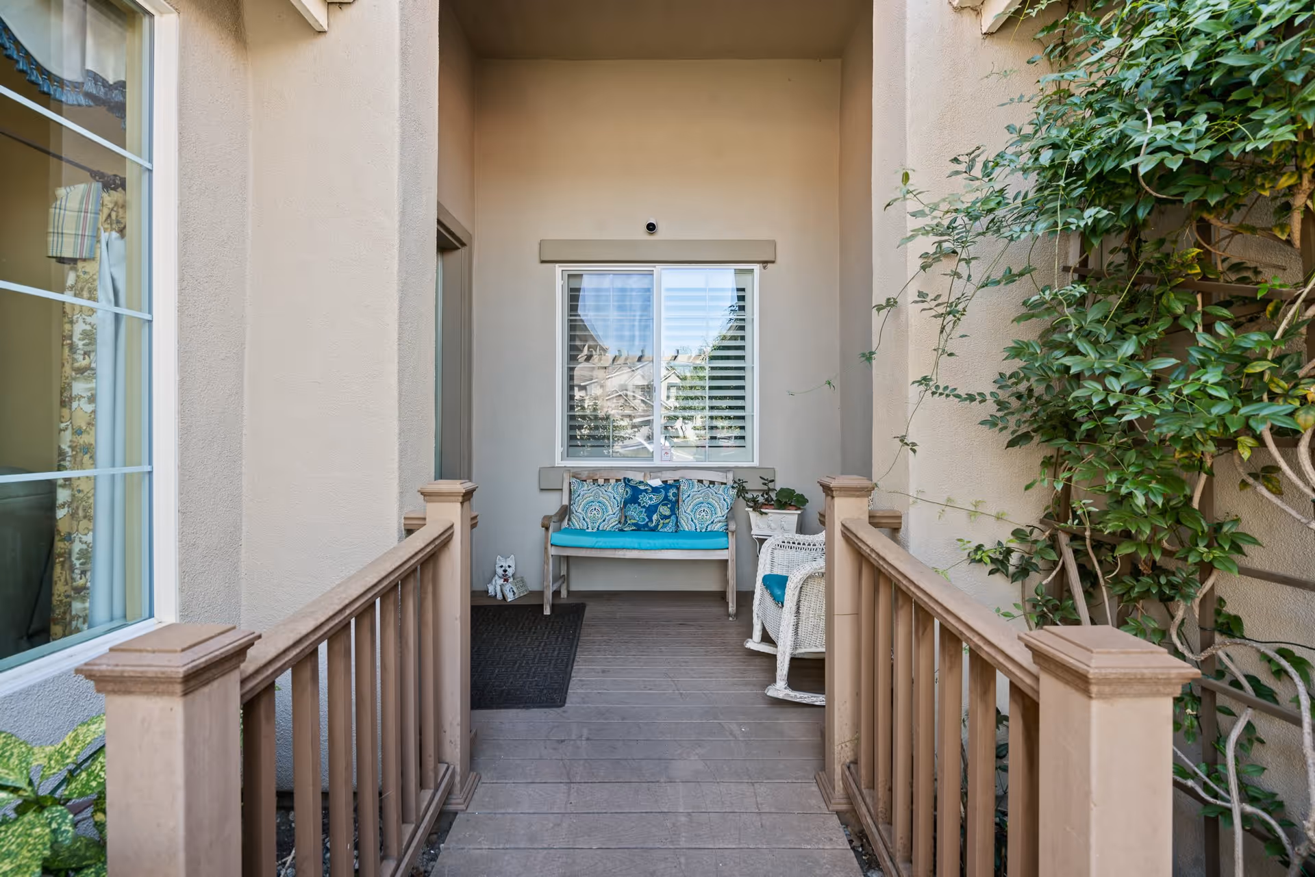 A covered outdoor porch area with a wooden bench adorned with blue and patterned cushions, a white wicker rocking chair with a blue cushion, a small white side table with a potted plant, and a small decorative dog statue near a black doormat. The porch is flanked by beige walls and wooden railings, with green leafy plants climbing on the right side.