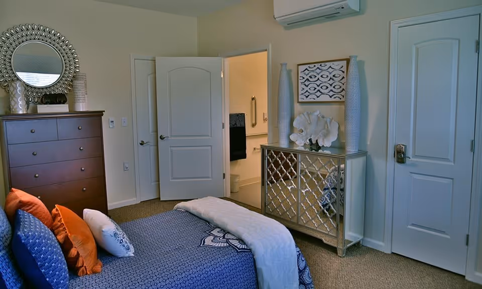 A bedroom with a bed covered in blue and white bedding and multiple pillows in blue, orange, and white. There is a dark wooden dresser with decorative items and a round mirror on the left side. A mirrored cabinet with two tall vases and a coral decoration is on the right side. Two white doors are visible, one slightly open showing a bathroom with grab bars and a towel rack.