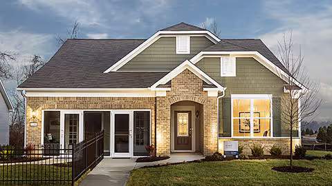 Exterior front view of a single-story house with a combination of brick and green siding, a dark shingled roof, a front door with sidelights, and large windows. The house is surrounded by a small lawn and a black metal fence on the left side.