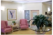 A nursing-home waiting area with two pink armchairs beside a small table, framed artwork on the wall, a glass door, and a glass-top table holding a large potted plant.