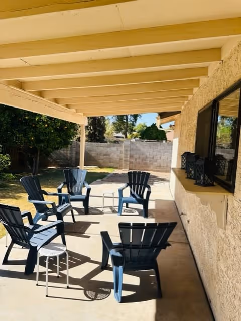 Outdoor covered patio area with six dark blue Adirondack chairs arranged in a circle on a concrete floor. A small white side table is also present. The patio is attached to a beige stucco building with a window and decorative lanterns on the windowsill. In the background, there is a grassy yard with trees and a cinder block wall.