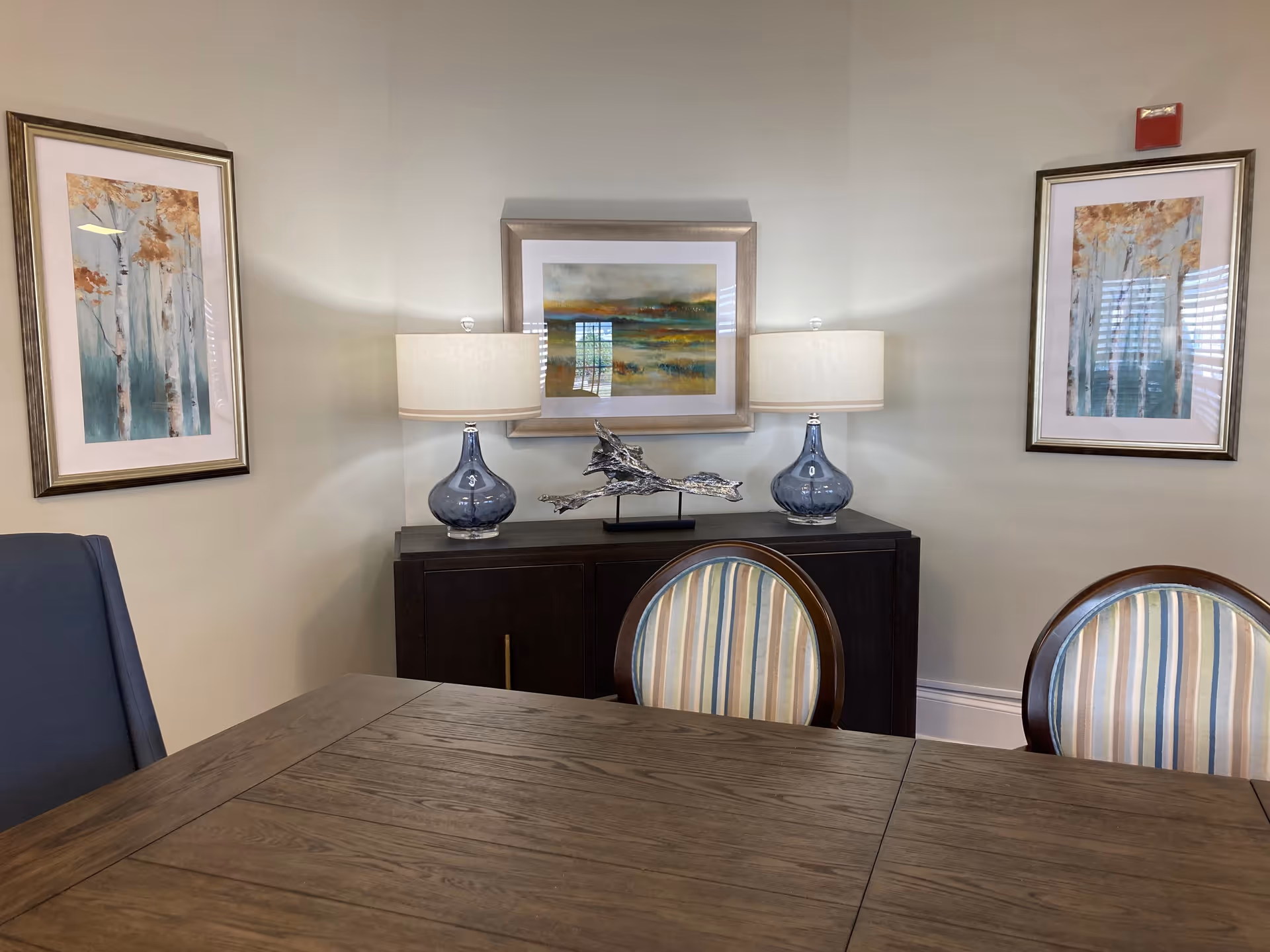 Dining room with a wooden table, striped chairs, a dark sideboard topped with blue lamps and decorative art on the walls.