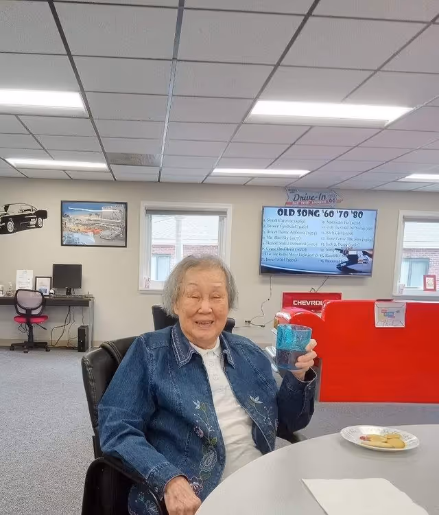 An elderly woman sitting at a round table in a common area, smiling and holding a blue cup. On the table are a plate with cookies and a napkin. The room has a gray carpet, a window, a TV screen displaying a list of old songs from the 60s, 70s, and 80s, and a red couch in the background. There is also a desk with a computer and chair along the wall.