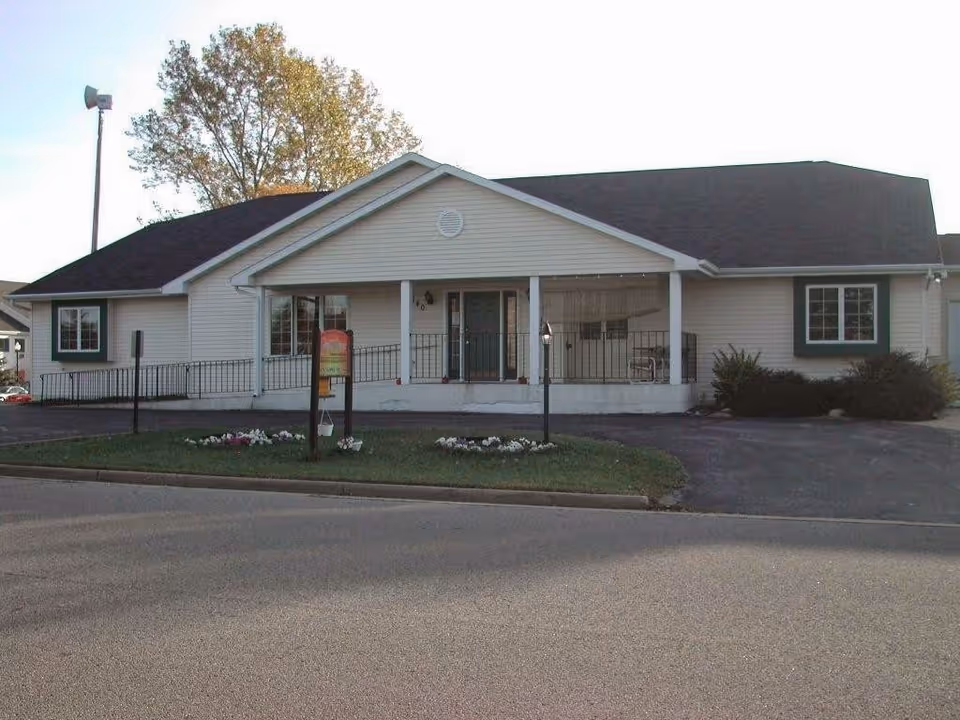Exterior view of a single-story building with beige siding and a dark roof, featuring a front porch with a railing and a ramp. There are two windows with green trim, a small garden with flowers in front, and a tree in the background.