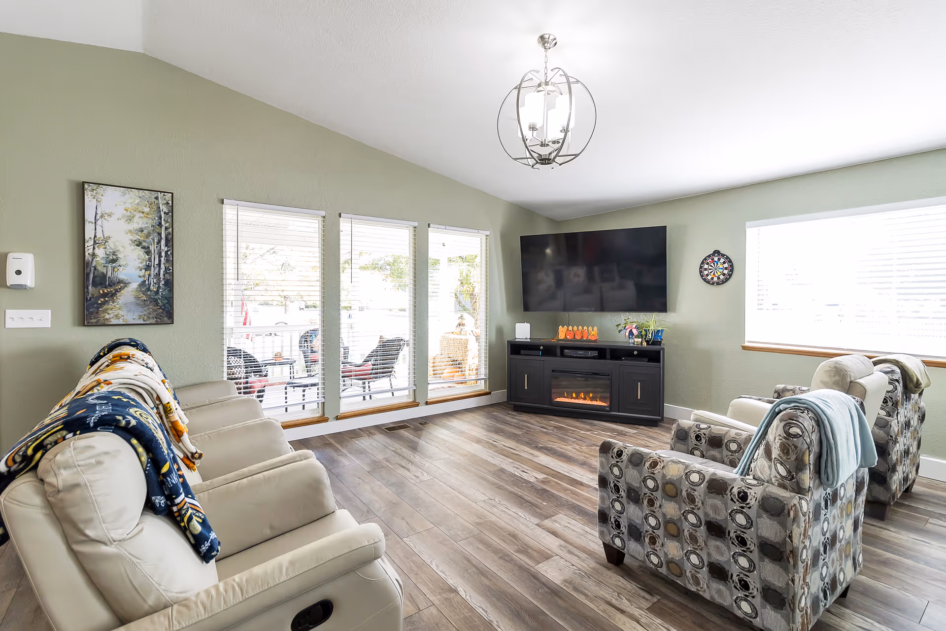 A bright and cozy living room with light green walls and wood flooring. The room features a large flat-screen TV mounted on the wall above a black electric fireplace. There are four comfortable chairs arranged facing the TV, two beige recliners with patterned blankets draped over them and two patterned armchairs with light blue blankets. Large windows with white blinds allow natural light to fill the space. A modern chandelier hangs from the ceiling, and a small dartboard is mounted on the wall next to the TV. Outside the windows, patio furniture is visible.