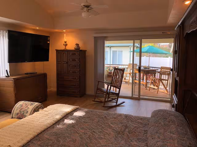 Bedroom with a bed in the foreground, wall-mounted TV and wooden dressers, and sliding glass doors opening to a patio with a table and umbrella.