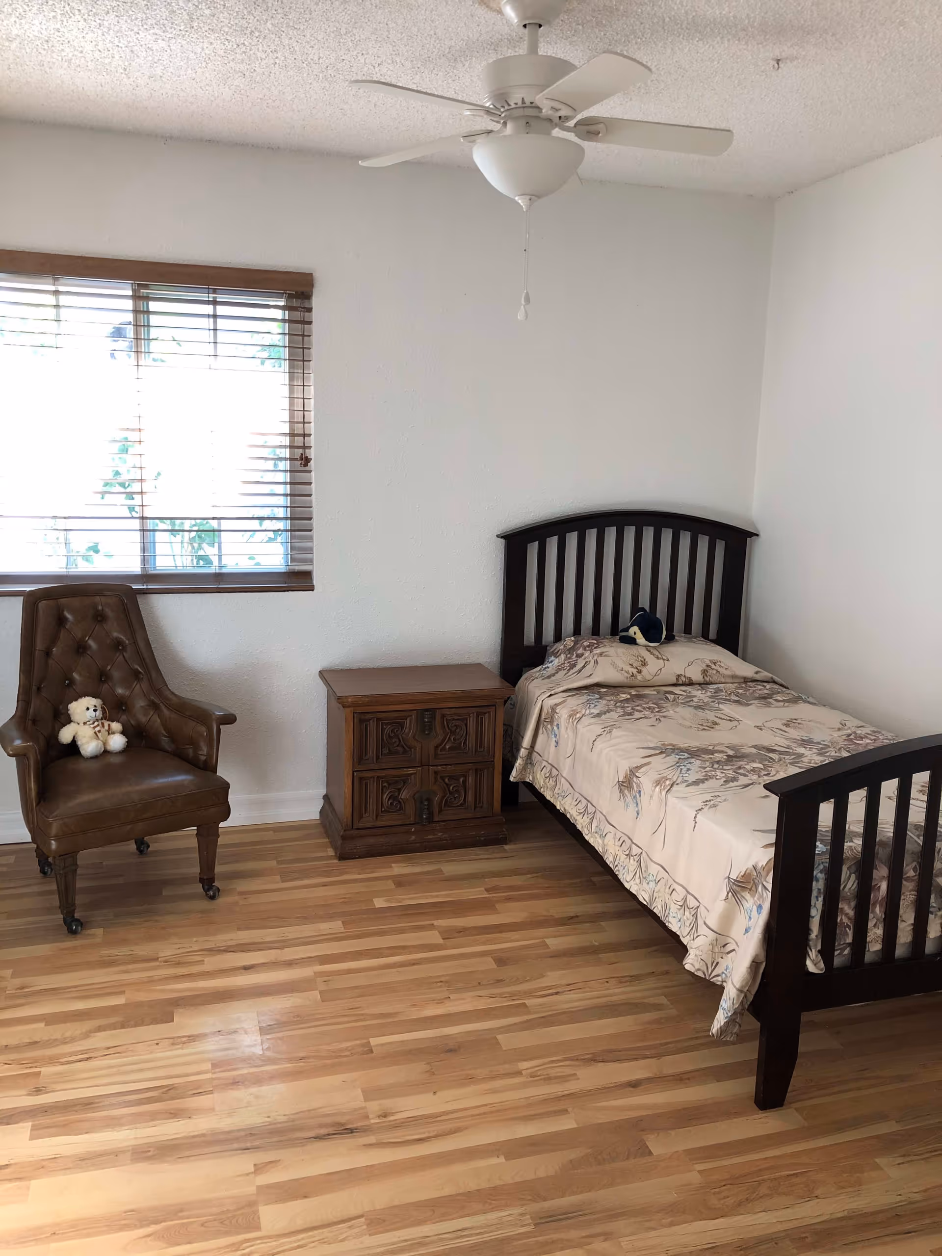 A simple bedroom with a single bed covered with a floral bedspread, a wooden nightstand, a brown leather armchair with a teddy bear on it, a window with wooden blinds, and a white ceiling fan.