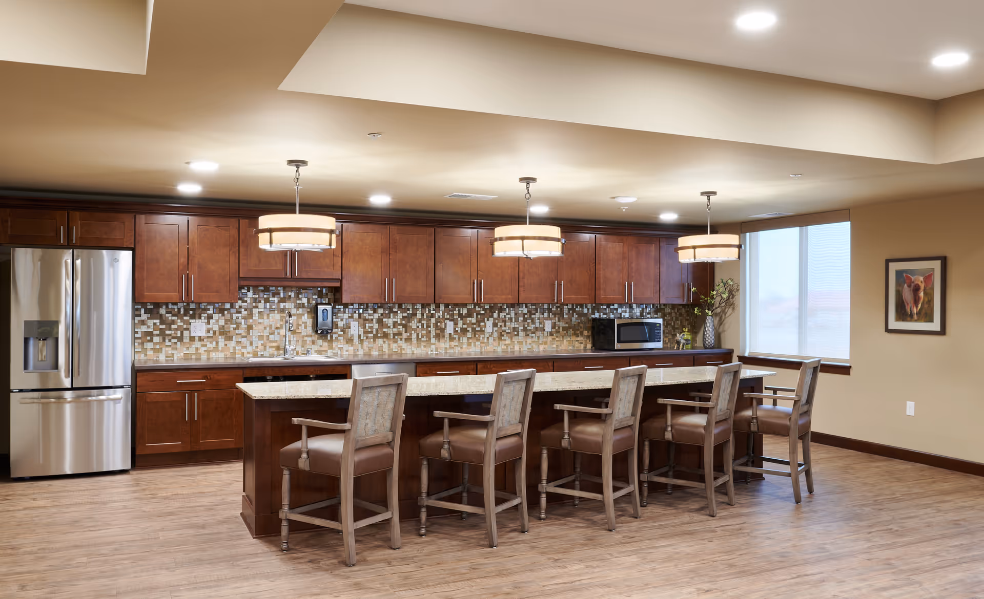 A spacious kitchen area with wooden cabinets, a mosaic tile backsplash, and a large island with six wooden chairs. The kitchen features a stainless steel refrigerator, a microwave, and three pendant lights hanging above the island. There is a window with blinds and a framed picture on the wall.