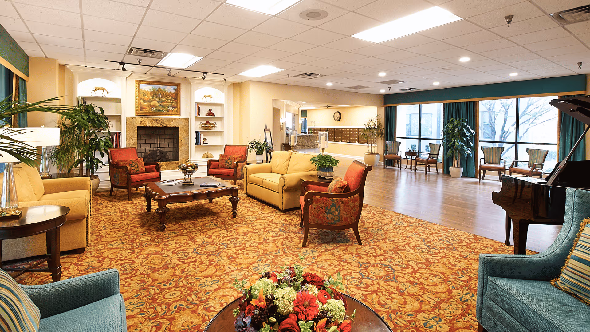 A spacious senior living facility common area with a patterned carpet, multiple upholstered chairs and sofas in warm colors, a wooden coffee table, a fireplace with built-in shelves, large windows with green curtains, several potted plants, and a grand piano on the right side.