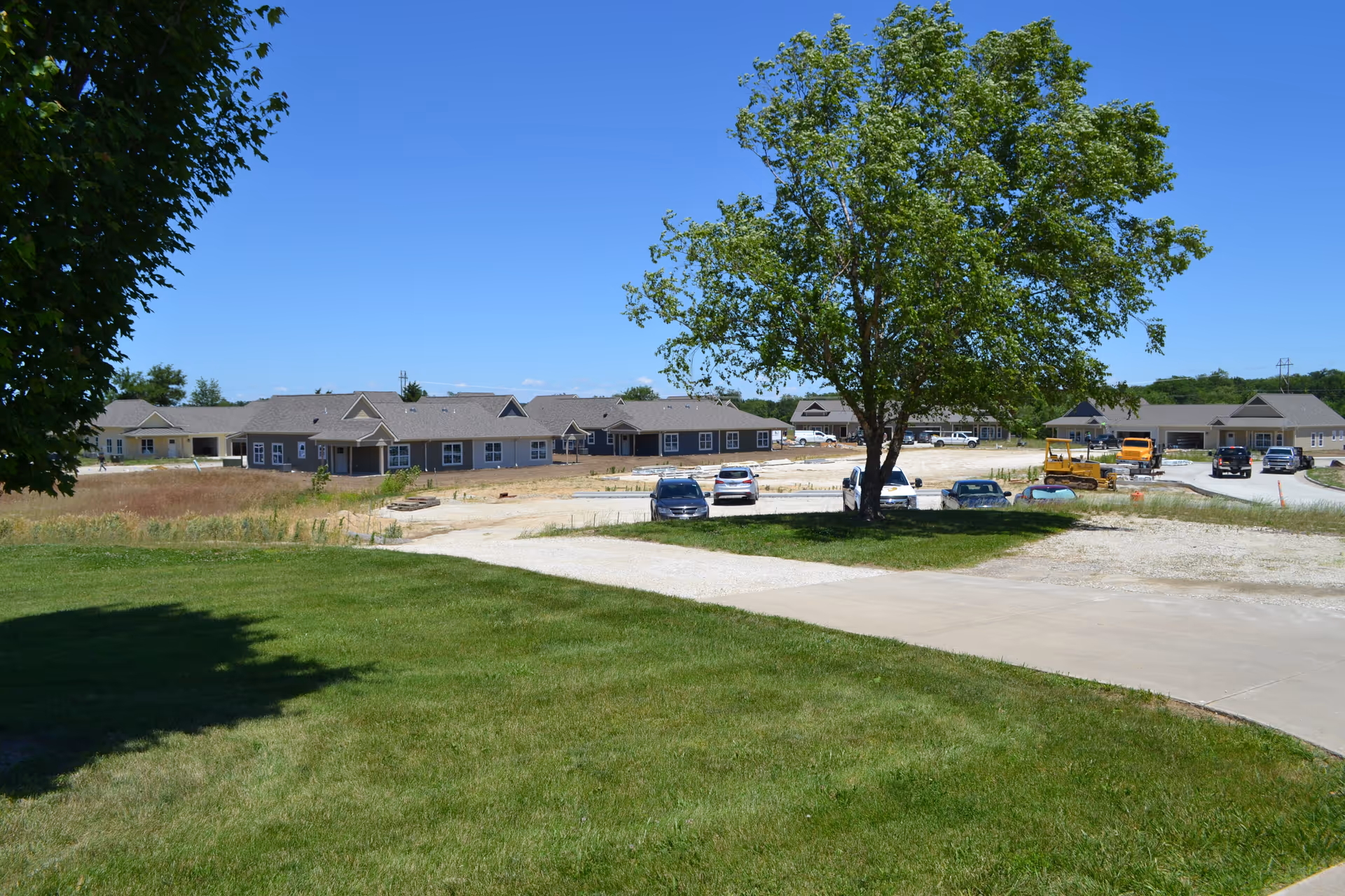View of a senior living facility under construction with multiple single-story buildings, a large tree in the center, several parked cars, and a clear blue sky.