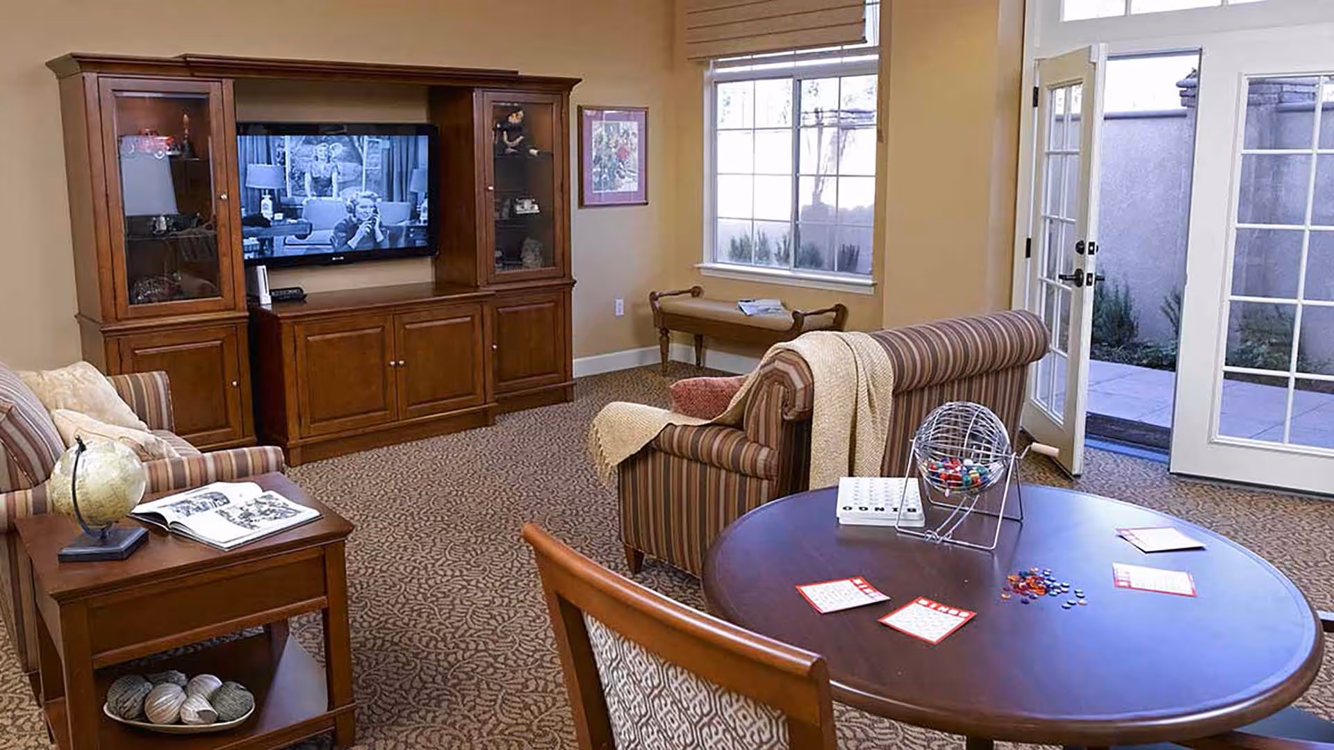 A cozy living room with a wooden entertainment center holding a TV playing a black and white show. The room features a striped armchair with a beige throw blanket, a wooden side table with a globe and an open book, a round table with bingo cards and a bingo cage, and a window and glass door leading to an outdoor patio.